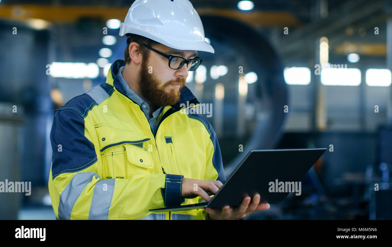 Industrial Engineer im harten Hut tragen Safety Jacket ist mit Laptop. Er arbeitet in der Schwerindustrie Fertigung Werk mit verschiedenen Metall Details Stockfoto