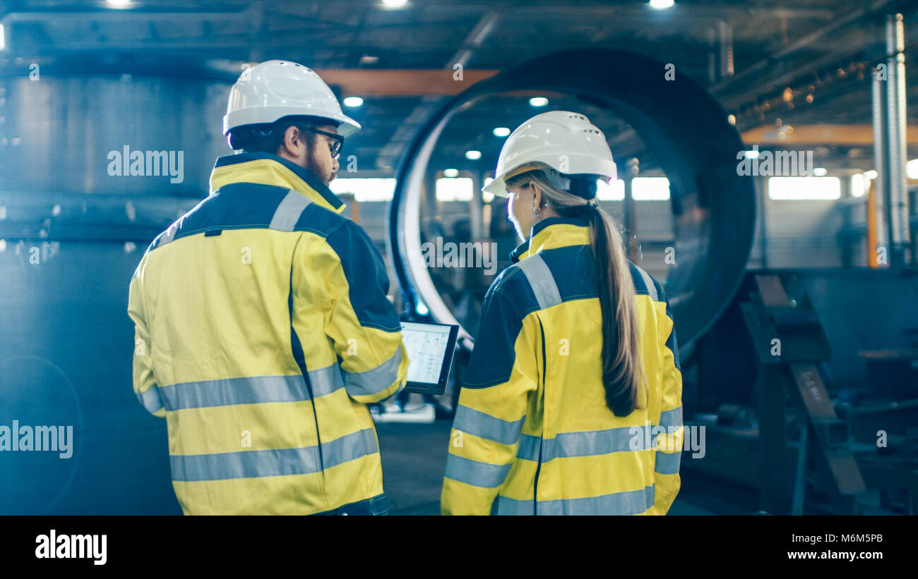 Männliche und weibliche Ingenieure mit Laptop und Diskussion während man durch Heavy Industry Manufacturing Factory. Sie harte Hüte tragen Stockfoto