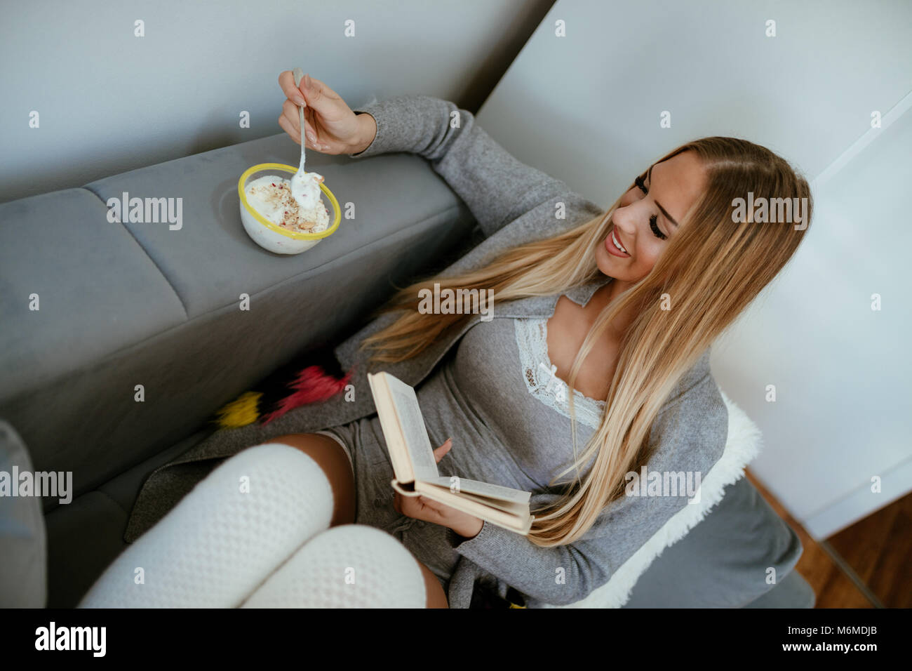 Attraktive lächelnde Mädchen auf dem Bett sitzen, gesundes Frühstück mit Müsli und Joghurt und Lesen Buch. Ansicht von oben. Stockfoto