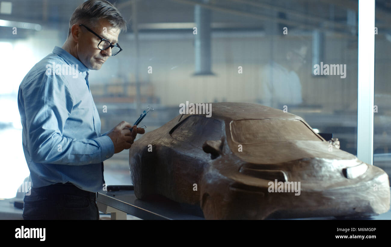 Chief Automotive Designer mit Rechen formt futuristischen Auto Modell aus Plastilin Ton. Er arbeitet in einem speziellen Studio In einem großen Auto Fabrik befindet. Stockfoto