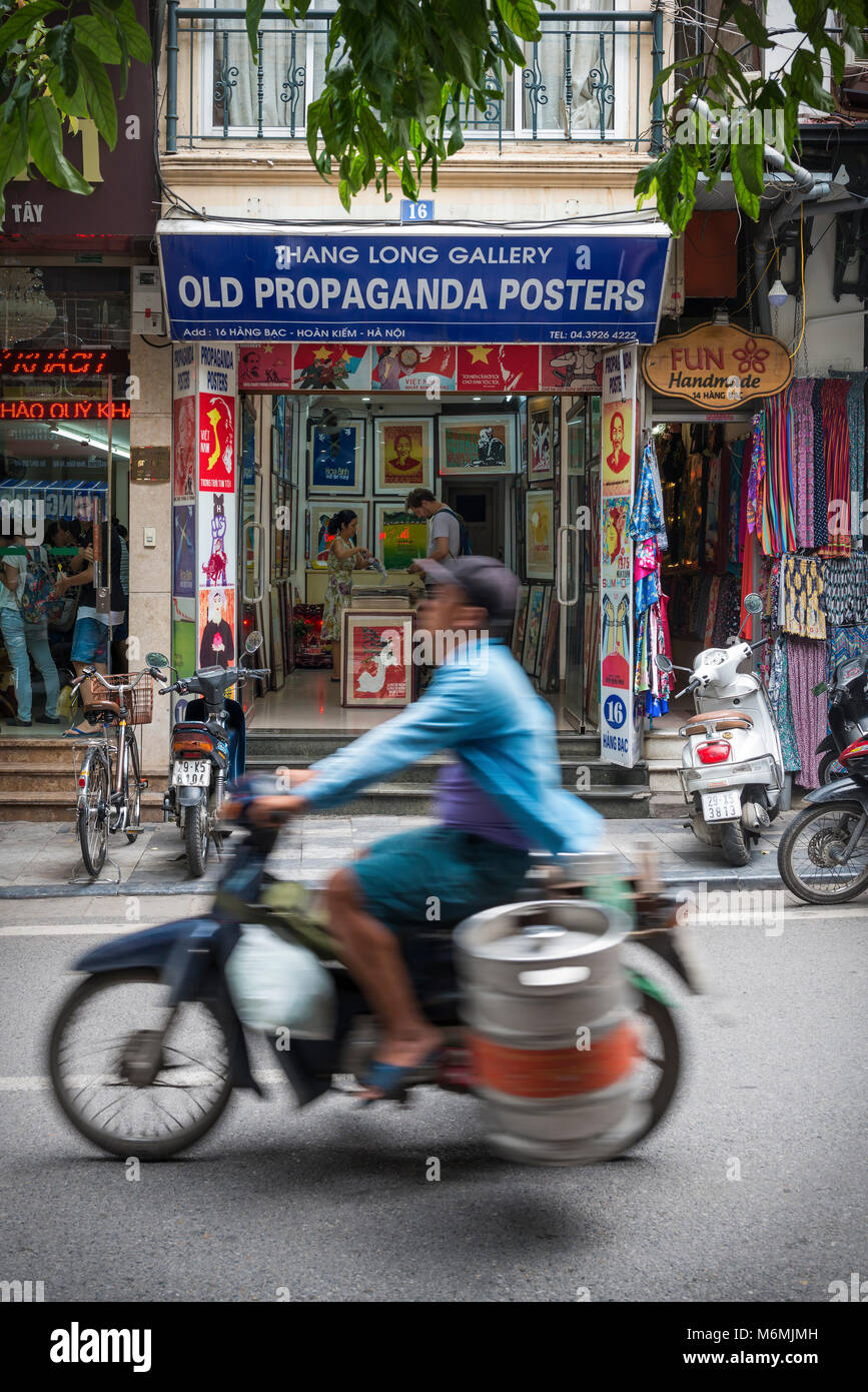 HANOI, VIETNAM - 14. Juli: Ein Mann transportiert ein Faß auf ein Moped vorbei an einem alten Propaganda Poster Shop in der Altstadt von Hanoi am 14 Juli, 2017 in Han Stockfoto