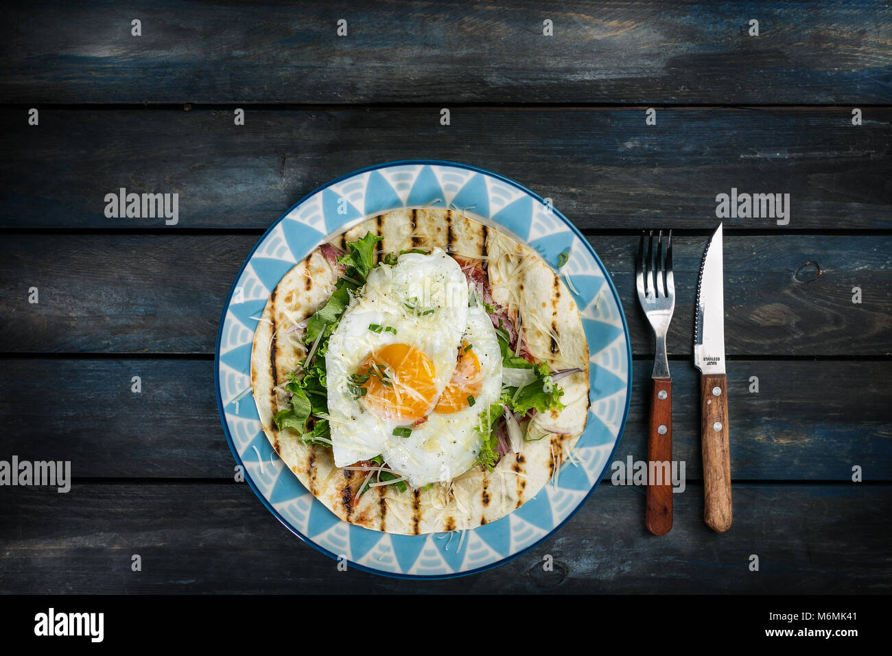Spiegeleier auf Mehl Tortilla mit grünem Salat und Käse. Nützliche Frühstück oder Mittagessen Idee. Gabel, Messer und schönen Teller auf einem farbigen Hintergrund aus Holz. Ansicht von oben Stockfoto