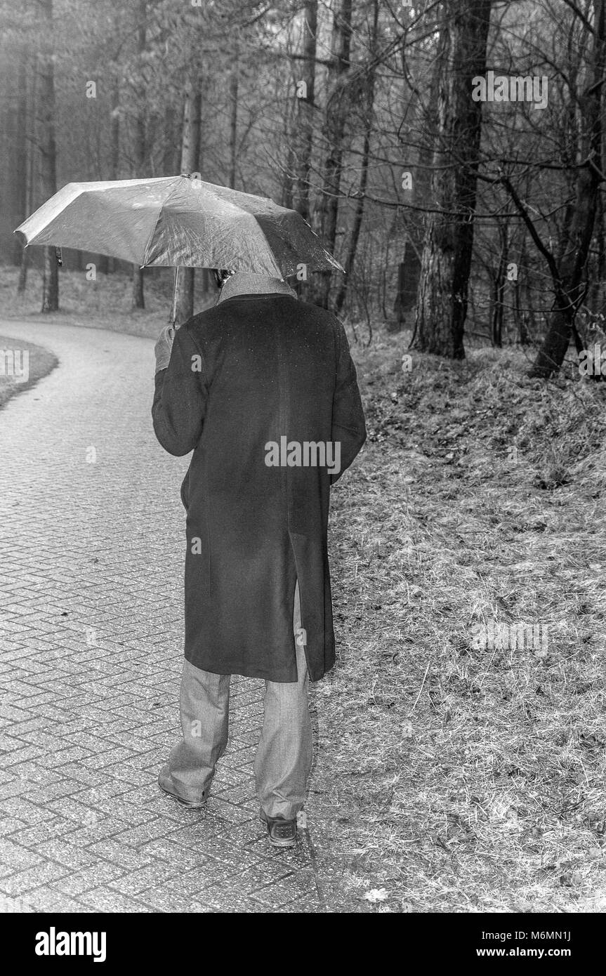 Schwarz und Weiß, Szene von einem Mann mit Regenmantel und Regenschirm Spaziergang im Wald Stockfoto