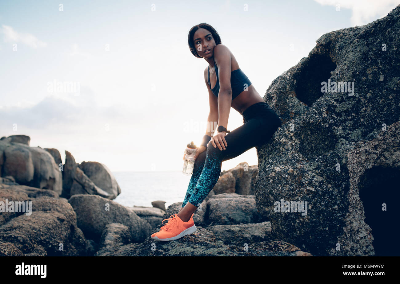 Fitness Frau mit einer Flasche Wasser ruhen über einen Felsen. Weibchen mit schlanken Körper Entspannung nach dem Training im Freien. Stockfoto
