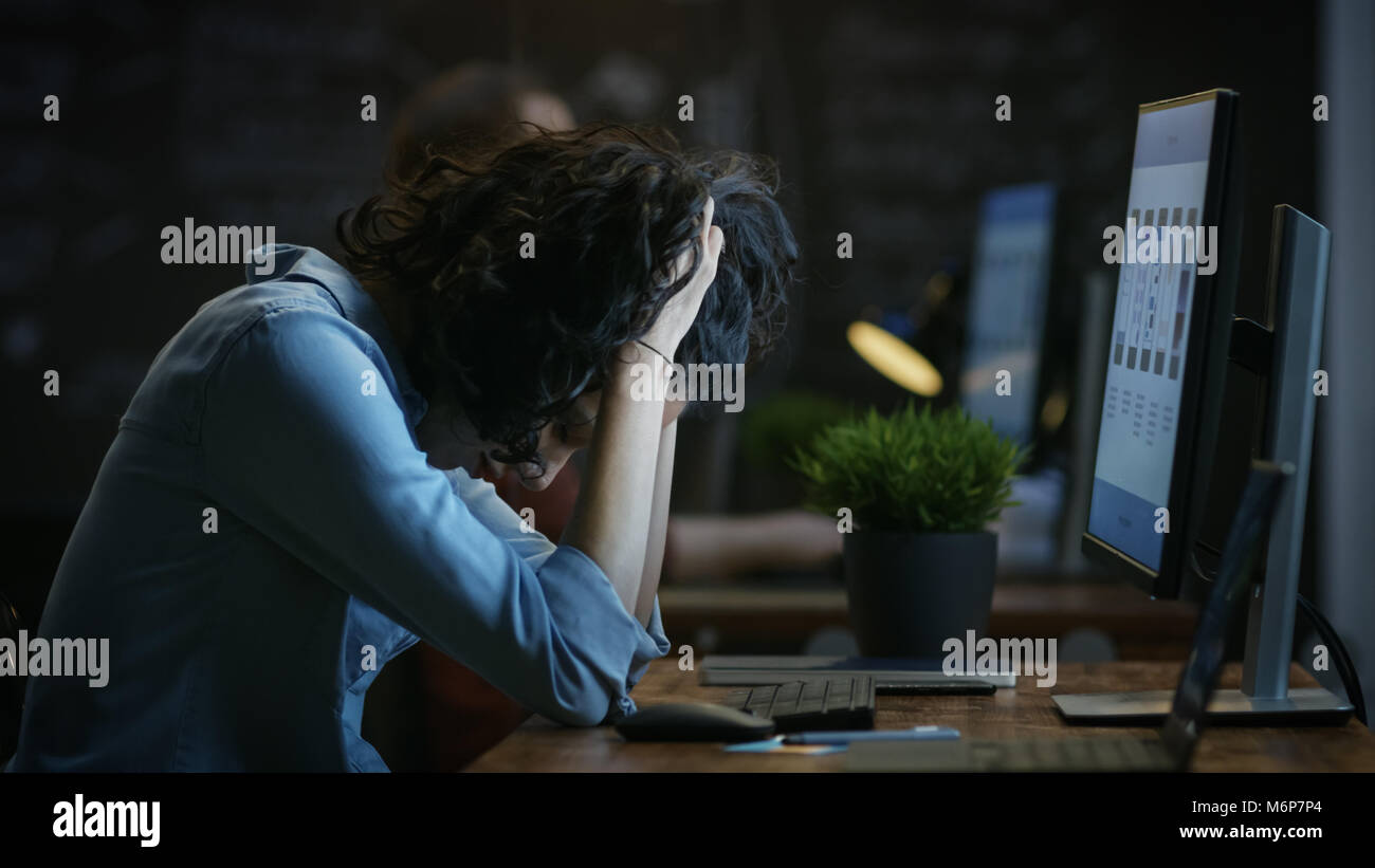 Müde, überarbeitete Weibliche Handy App Designer hält Ihren Kopf in die Hände während der Arbeit auf einem PC. Im Hintergrund kreative Büro. Stockfoto