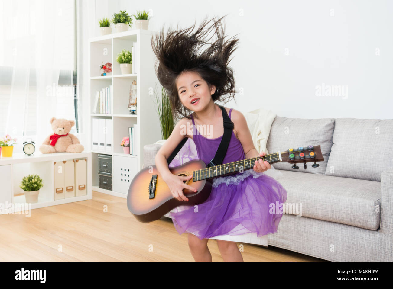 Kleine Mädchen Tochter Holding eine hölzerne Gitarre aufregend springt vom Sofa aus. Ihr Haar in die Luft. Freudige Konzept zu spielen. Stockfoto