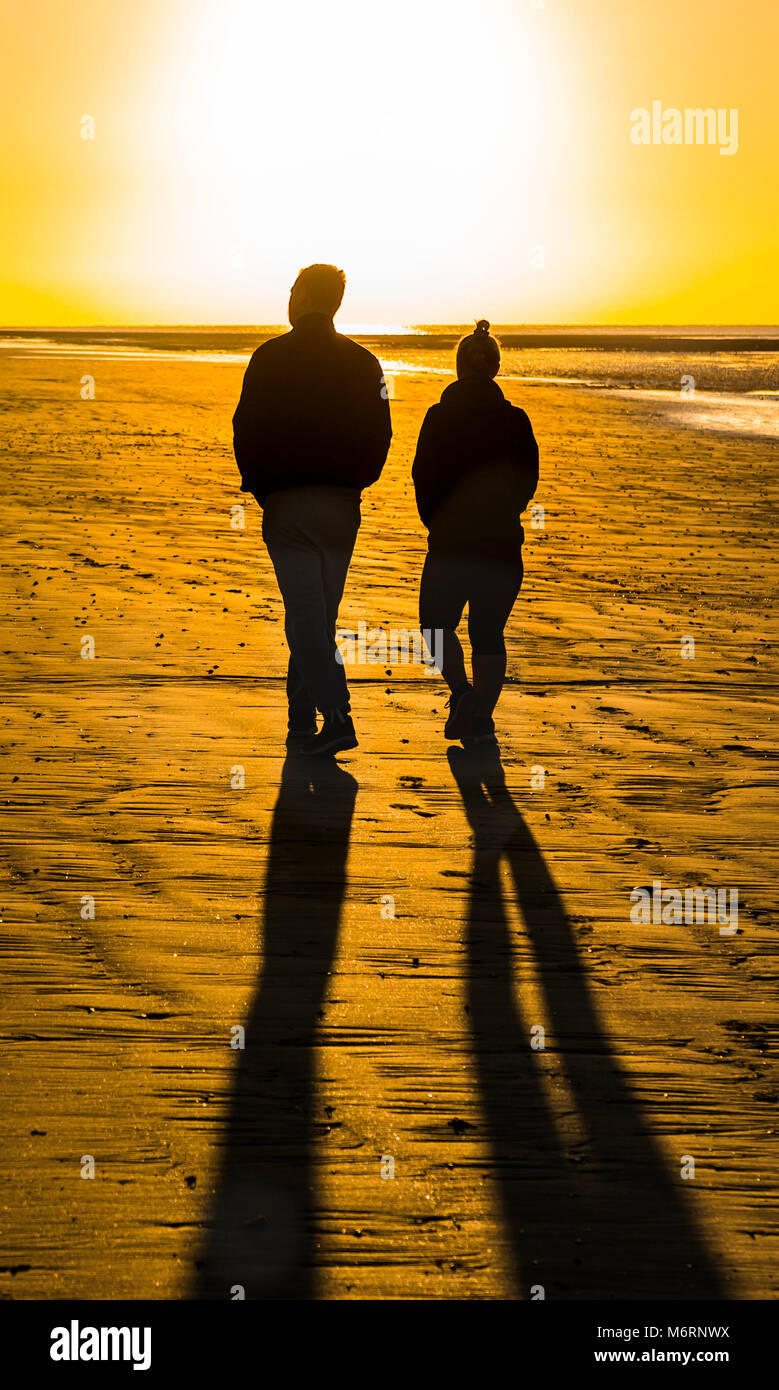 Ein paar Menschen zu Fuß auf einen Sandstrand am Morgen Richtung der aufgehenden Sonne, die wirft lange Schatten. Stockfoto