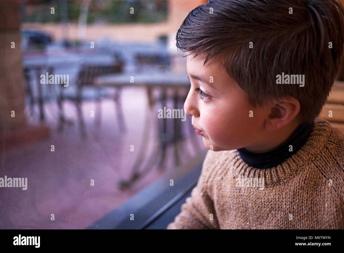 Gemischte Rasse asiatischen Kaukasischen kleiner Junge mit braunen Haar braun Pullover blickt ein Fenster auf einen Regentag und Gefühl Heimweh Stockfoto