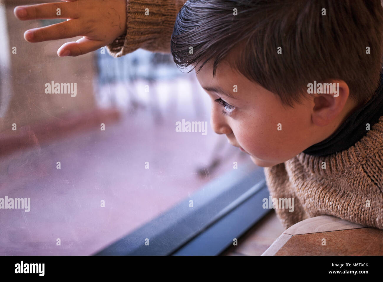 Gemischte Rasse asiatischen Kaukasischen kleiner Junge mit braunen Haar braun Pullover blickt ein Fenster auf einen Regentag und Gefühl Heimweh Stockfoto