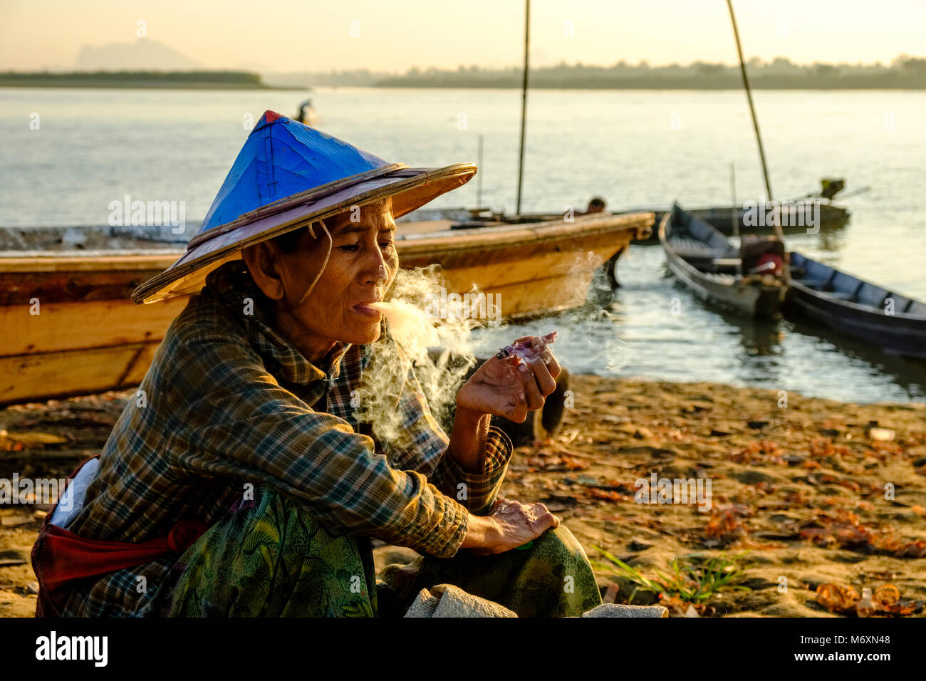 Eine Frau ist Rauchen eine Cheerot, einer lokalen Zigarre, Holz- Fischer Boote sind dicht am Ufer des Saluen Thanlyin Fluss Stockfoto