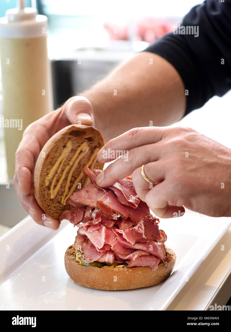 Der Küchenchef bereitet eine Pastrami Sandwich auf einem getoasteten Burgerbrötchen Schichtung der Reife würzige geräuchertem Rindfleisch Schichten in der Nähe zu sehen, der seine Hände Stockfoto