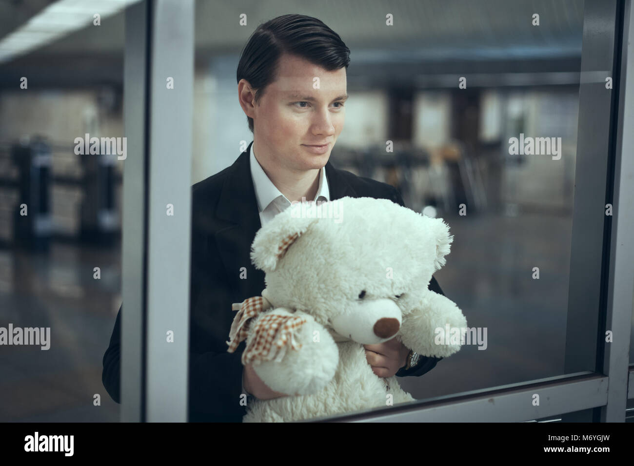 Junger Mann schaut aus dem Fenster und lächelt. Er hat einen Teddybär in der Hand Stockfoto