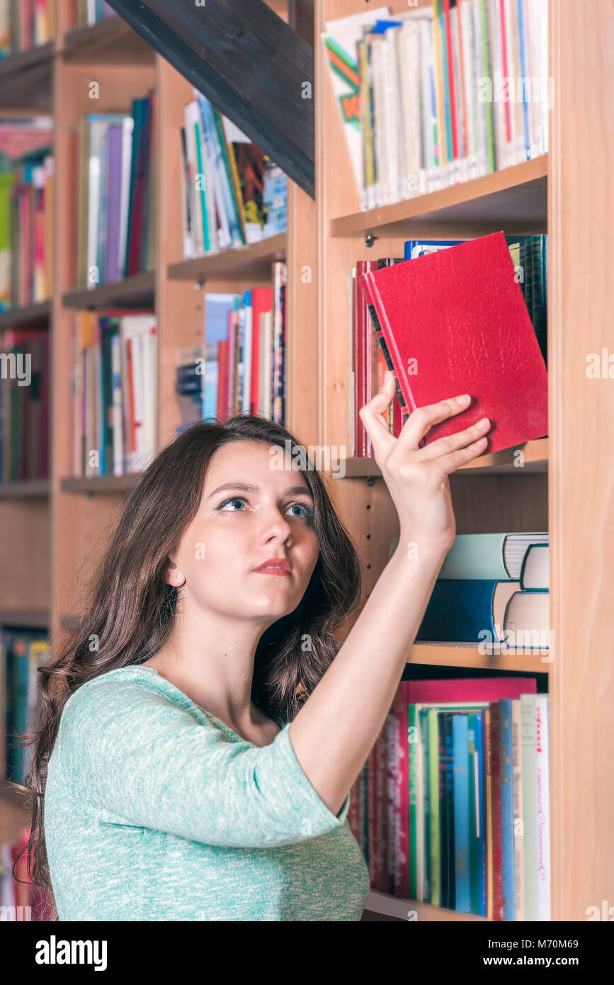Junge schöne Frau Auswahl ein rotes Buch aus einer Bibliothek. Ein Konzept für Bildung oder Freizeitaktivitäten. Stockfoto