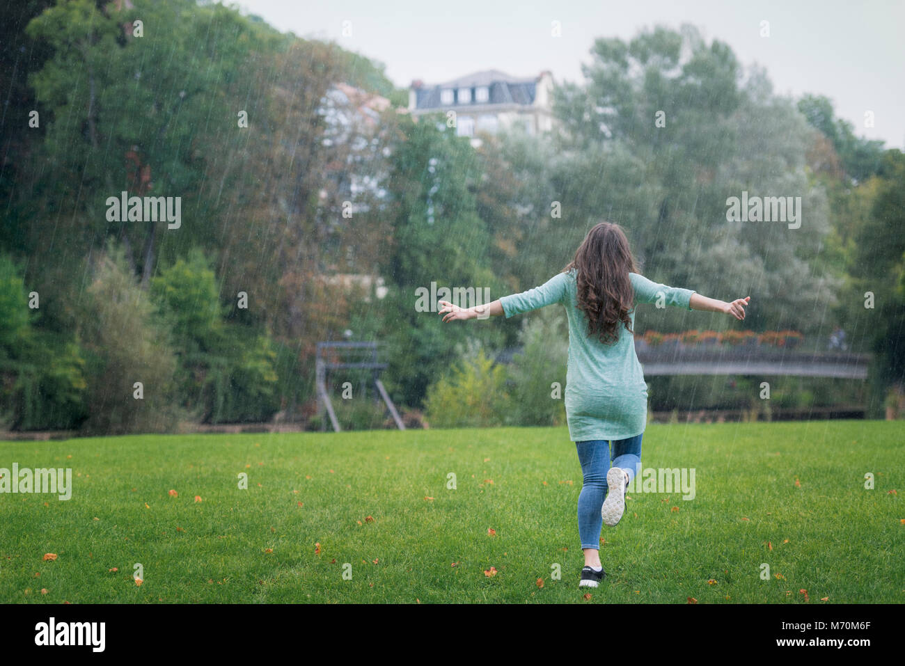 Brünette junge Frau gekleidet, casual, gerne Laufen im Regen, die Arme weit ausgebreitet, von grünem Gras und Bäumen umgeben, in Deutschland. Stockfoto