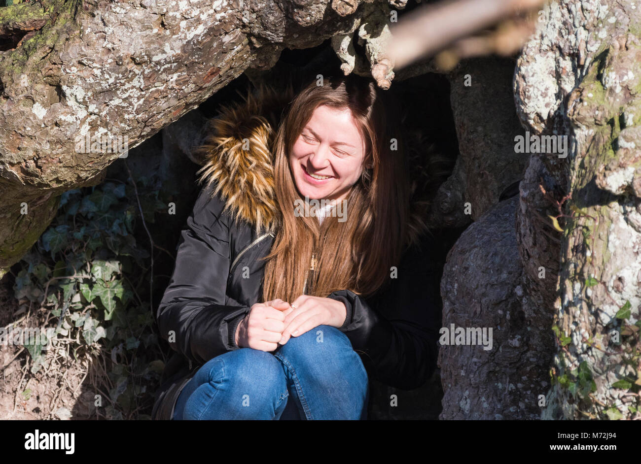 Hübsche junge Frau sitzt draußen unter einem Baum trägt einen Mantel, an einem kalten Tag im Winter mit Sonne im Gesicht. Stockfoto