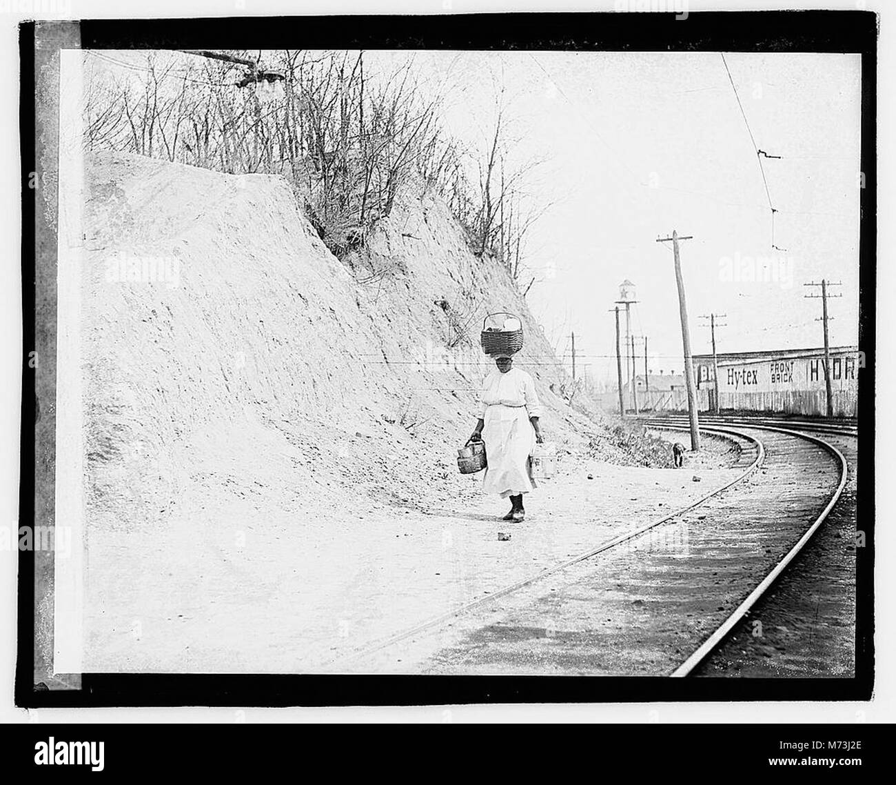 Dieses Foto zeigt eine Afroamerikanerin, die mit einem Korb auf dem Kopf in der Nähe von Eisenbahnschienen spaziert und das tägliche Leben und die Arbeit der Afroamerikaner im frühen 20. Jahrhundert festnimmt. Stockfoto