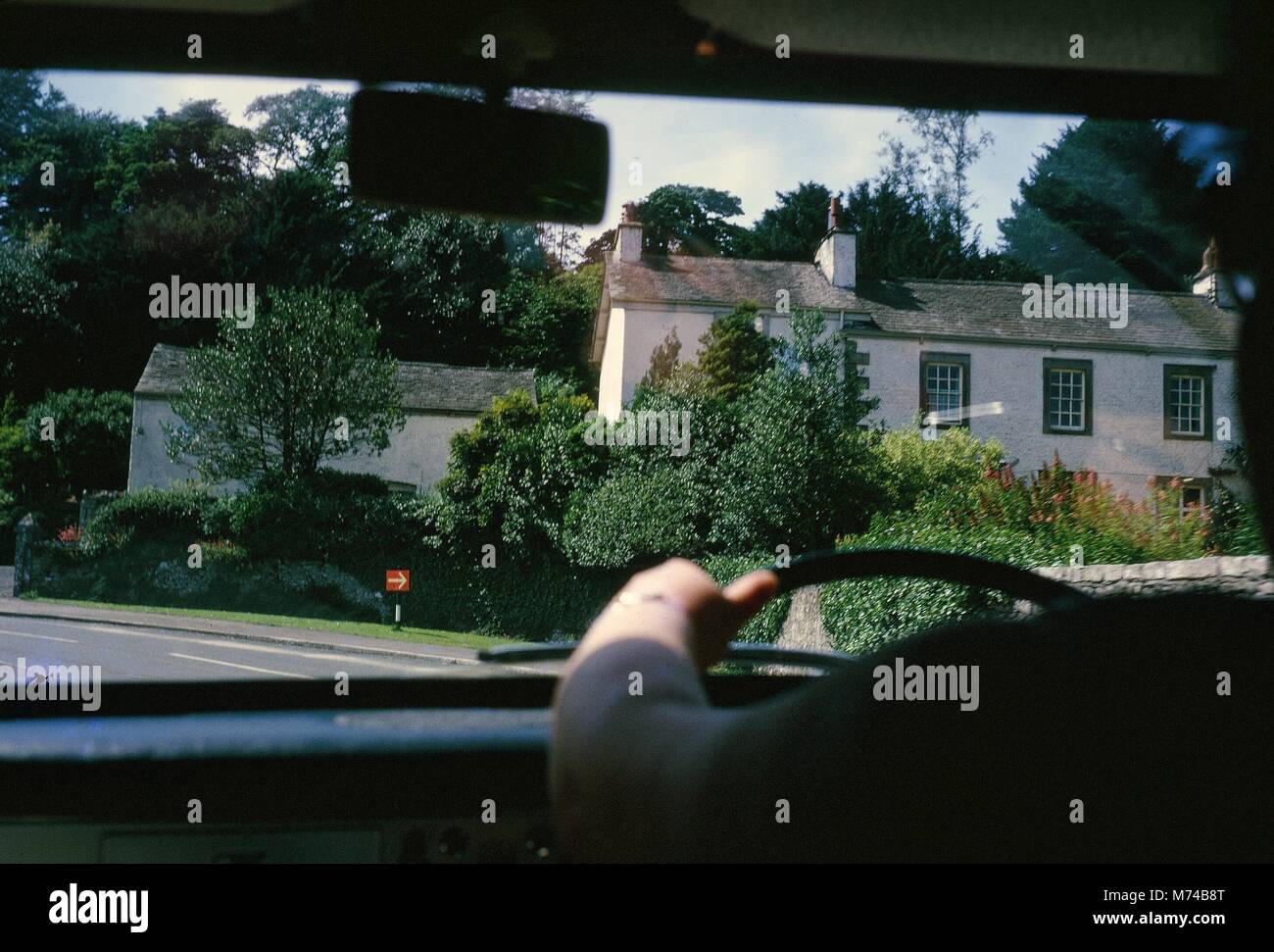 Ein Haus mit Bäumen umgeben ist teilweise verdeckt durch die Hände des Fahrers, Lenkrad und Armaturenbrett des Taxi, 1965. () Stockfoto
