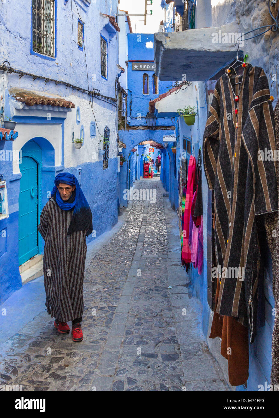 Ein lokaler Mann in traditioneller Kleidung Kleidung Wanderungen entlang der bunten engen gepflasterten Straße in die blaue Stadt Chefchaouen, Marokko. Djebella auflegen. Stockfoto