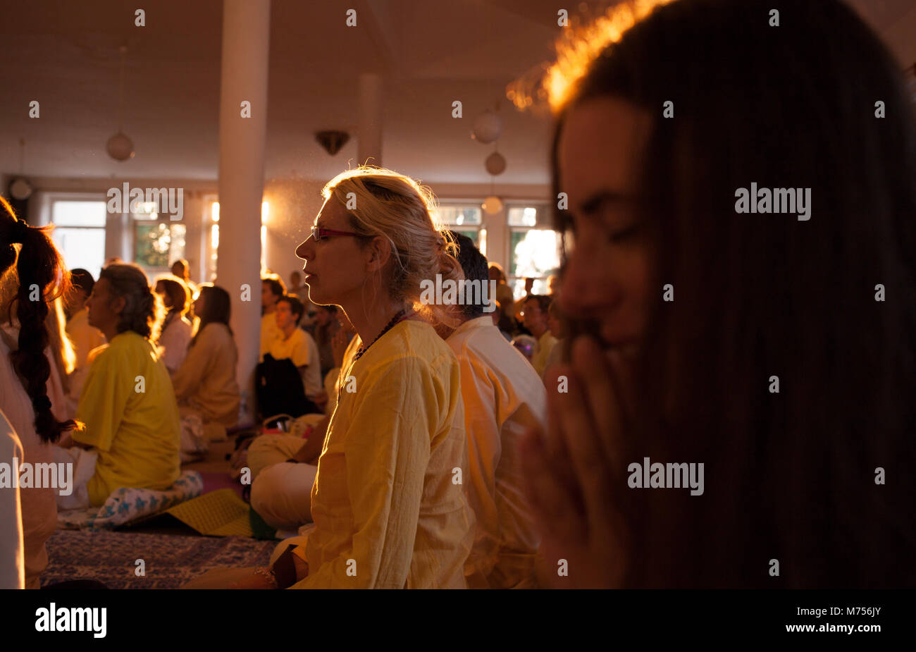 Eine Halle voller Menschen, die beten mit gefalteten Händen und geschlossenen Augen, während die Sonne durch ein Windows scheint während des Sonnenuntergangs. Leute, Sommer yoga Stockfoto