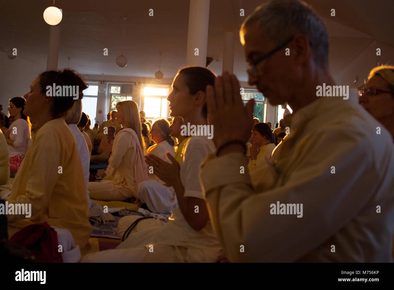 Eine Halle voller Menschen, die beten mit gefalteten Händen und geschlossenen Augen, während die Sonne durch ein Windows scheint während des Sonnenuntergangs. Leute, Sommer yoga Stockfoto