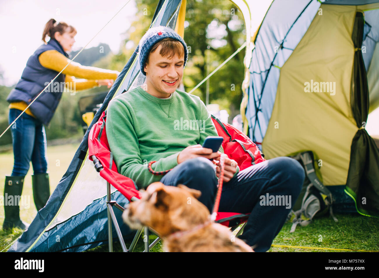 Teenager sitzt in einem camping Stuhl durch seine Hütte, in seinem smart phone verfasst, während seine Mamma stellt das Zelt. Stockfoto