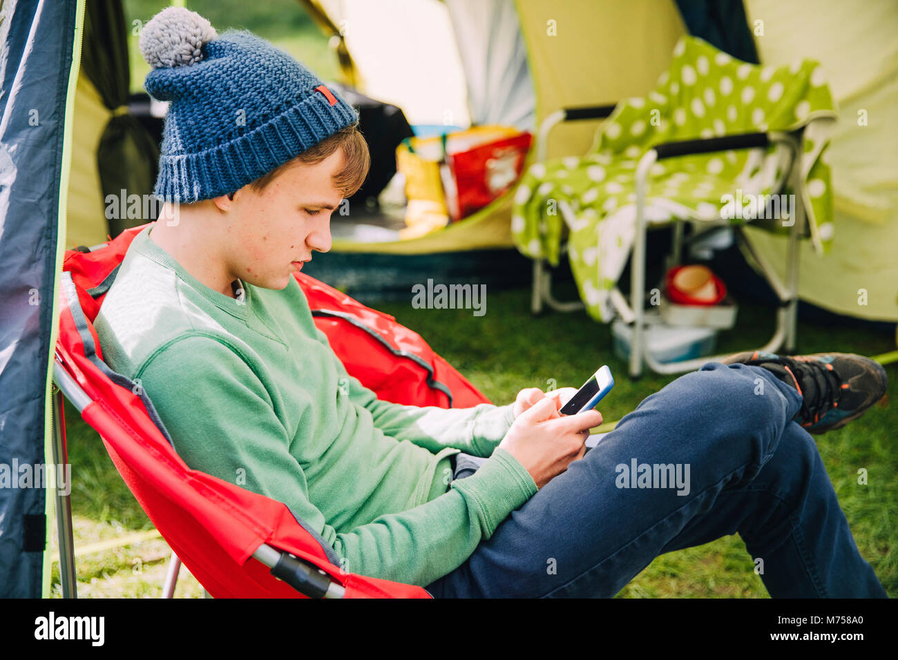 Teenager sitzt in einem camping Stuhl durch seine Hütte, in seinem smart phone verfasst. Stockfoto