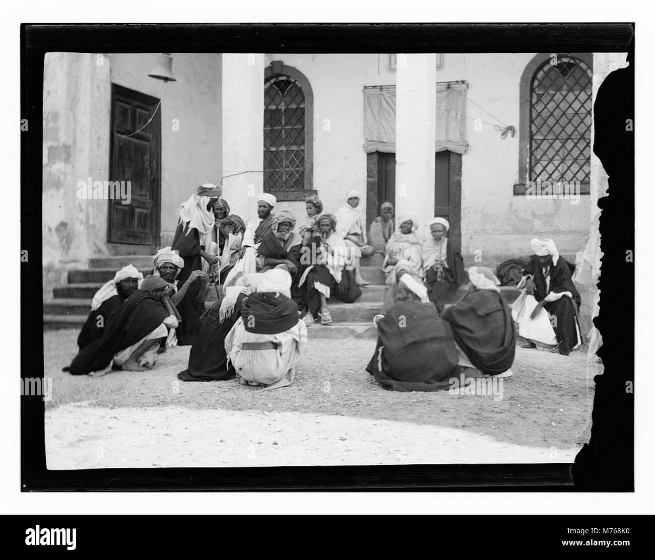 Ein Foto einer Gruppe von Männern, die auf den Stufen vor einem Gebäude sitzen und einen Moment der sozialen Interaktion oder der Freizeit in einer städtischen Umgebung festhalten. Stockfoto