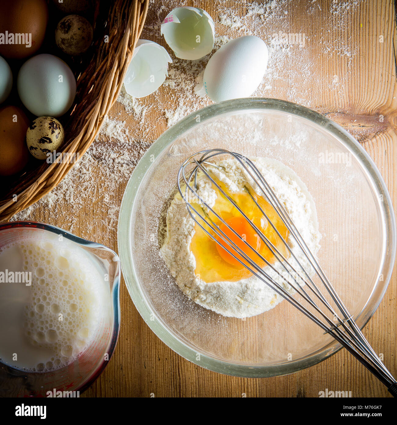 Overhead shot von Ei und Mehl in Schüssel mit Korb mit Eiern und kanne Milch zu einer Seite Stockfoto