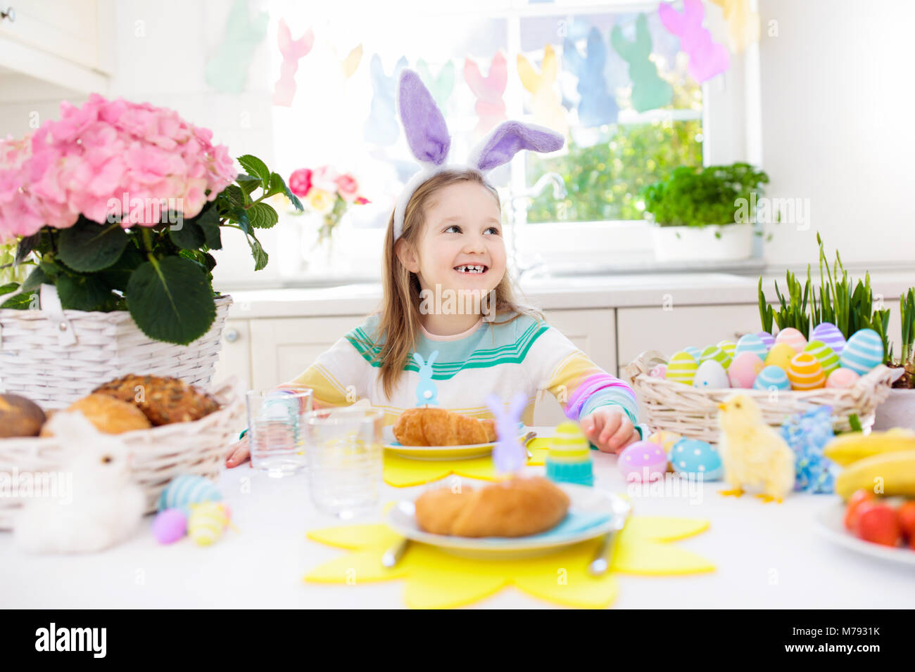 Familie Osterfrühstück. Kind mit Hasenohren an dekorierten Tisch Korb mit Eiern, Küken und Kaninchen am Ostermorgen. Ostereiersuche und festliche Mahlzeit für Stockfoto