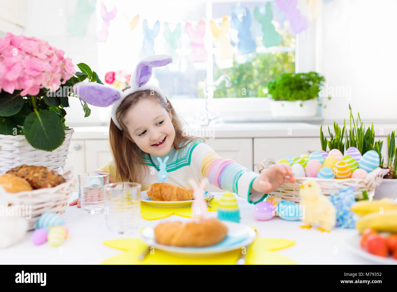 Familie Osterfrühstück. Kind mit Hasenohren an dekorierten Tisch Korb mit Eiern, Küken und Kaninchen am Ostermorgen. Ostereiersuche und festliche Mahlzeit für Stockfoto