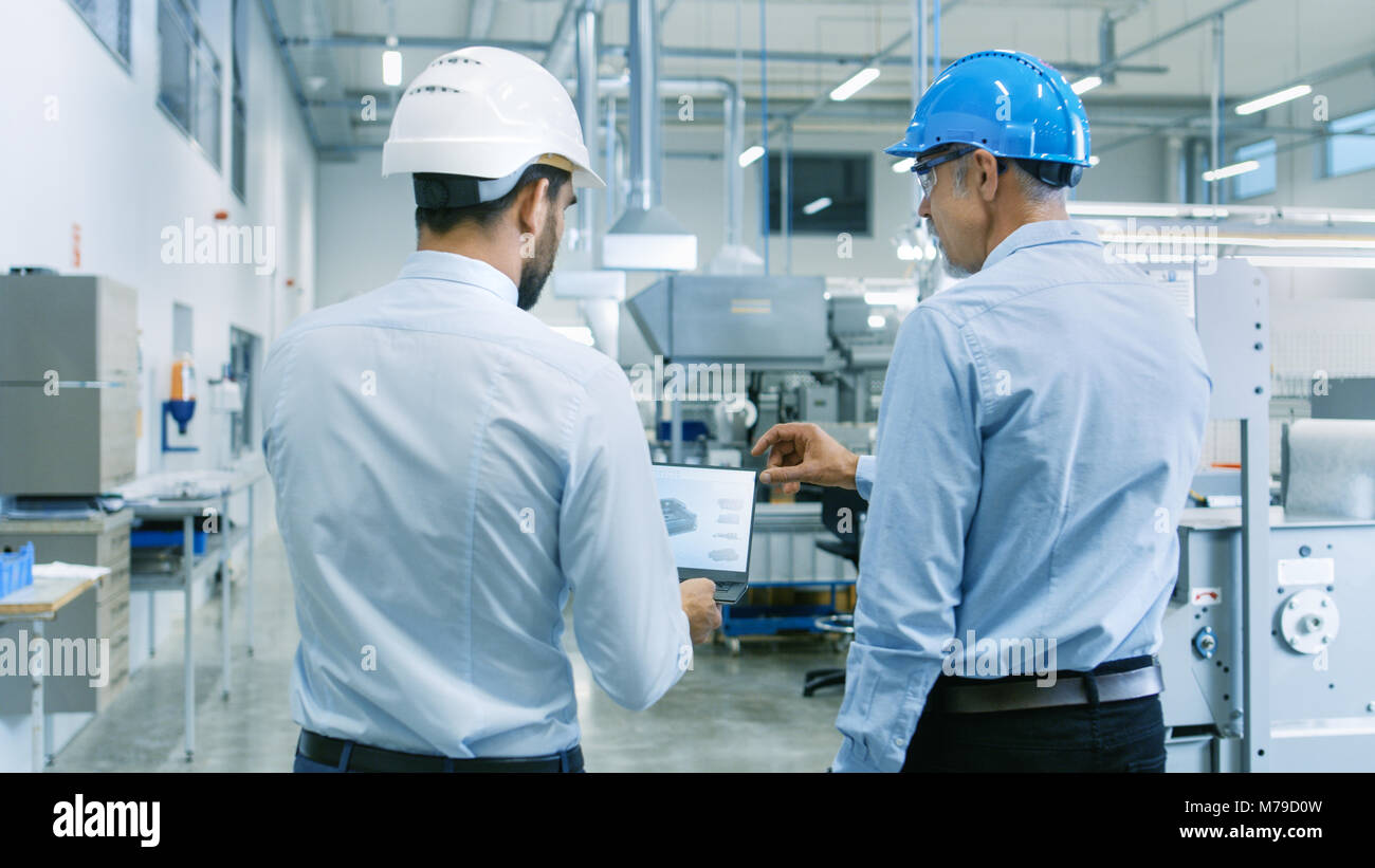 Rückansicht der Leiter des Projekts hält Laptop und Diskutieren Produkt Details mit Chief Engineer, während Sie durch moderne Fabrik gehen. Stockfoto