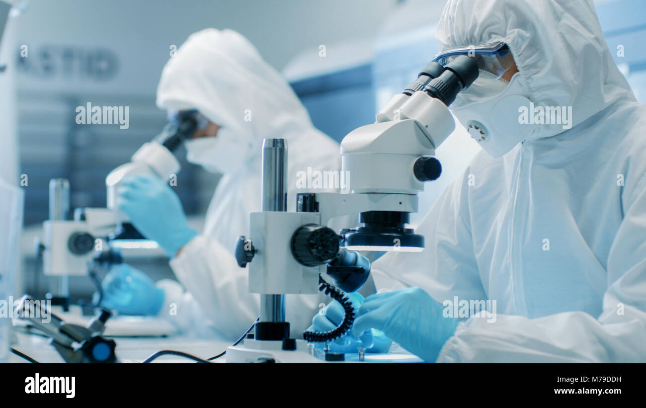 Zwei Ingenieure/Wissenschaftler/Techniker in steriler Reinraum Anzüge mit Mikroskopen für Komponente Anpassung und Forschung. Sie arbeiten in einem modernen Labor Stockfoto
