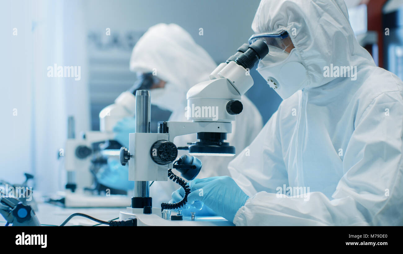 Zwei Ingenieure/Wissenschaftler/Techniker in steriler Reinraum Anzüge mit Mikroskopen für Komponente Anpassung und Forschung. Sie arbeiten in einem modernen Labor Stockfoto