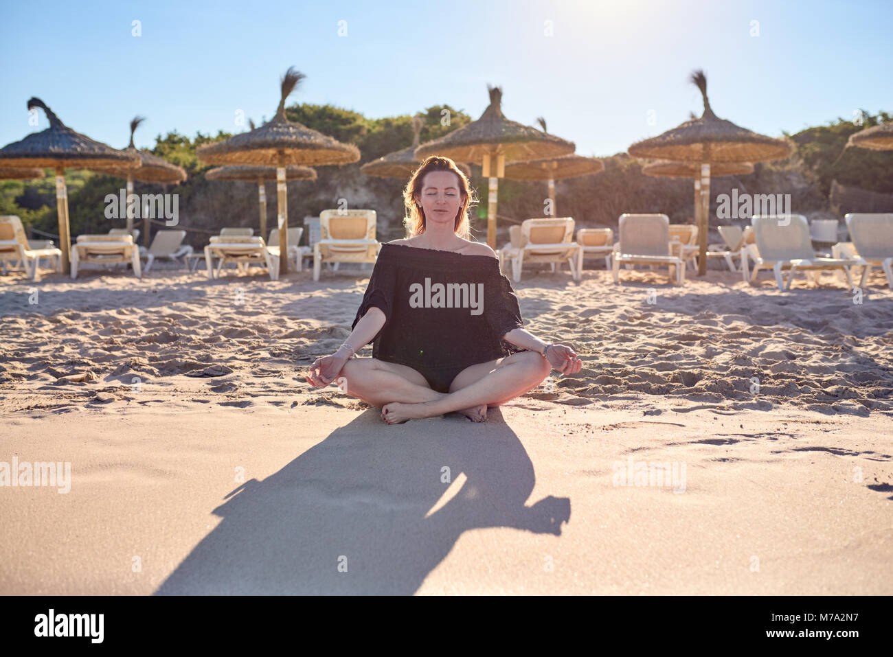 Attraktive barfuß Frau mit einem heiteren Ausdruck Meditation am Strand mit überkreuzten Beinen auf dem Sand Gegenlicht der Sonne vor dem Hintergrund der r Stockfoto