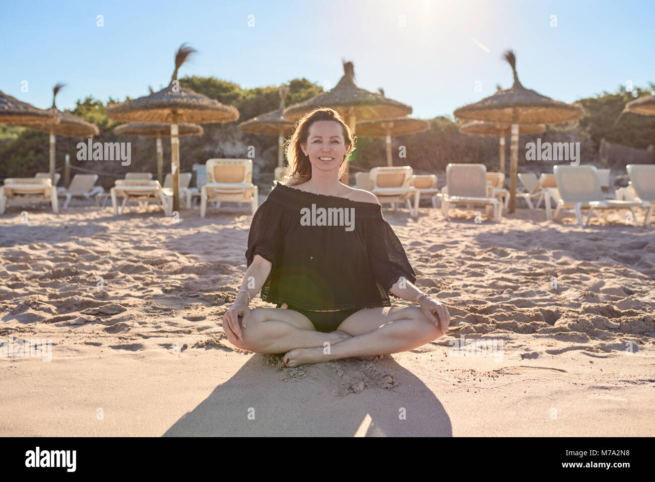 Lächelnd gesund barfuß Frau sitzt im Schneidersitz auf dem Strand, bereitet sie vor dem Hintergrund der Resort Sonnenschirme am Strand von t zu meditieren, mit Hintergrundbeleuchtung Stockfoto