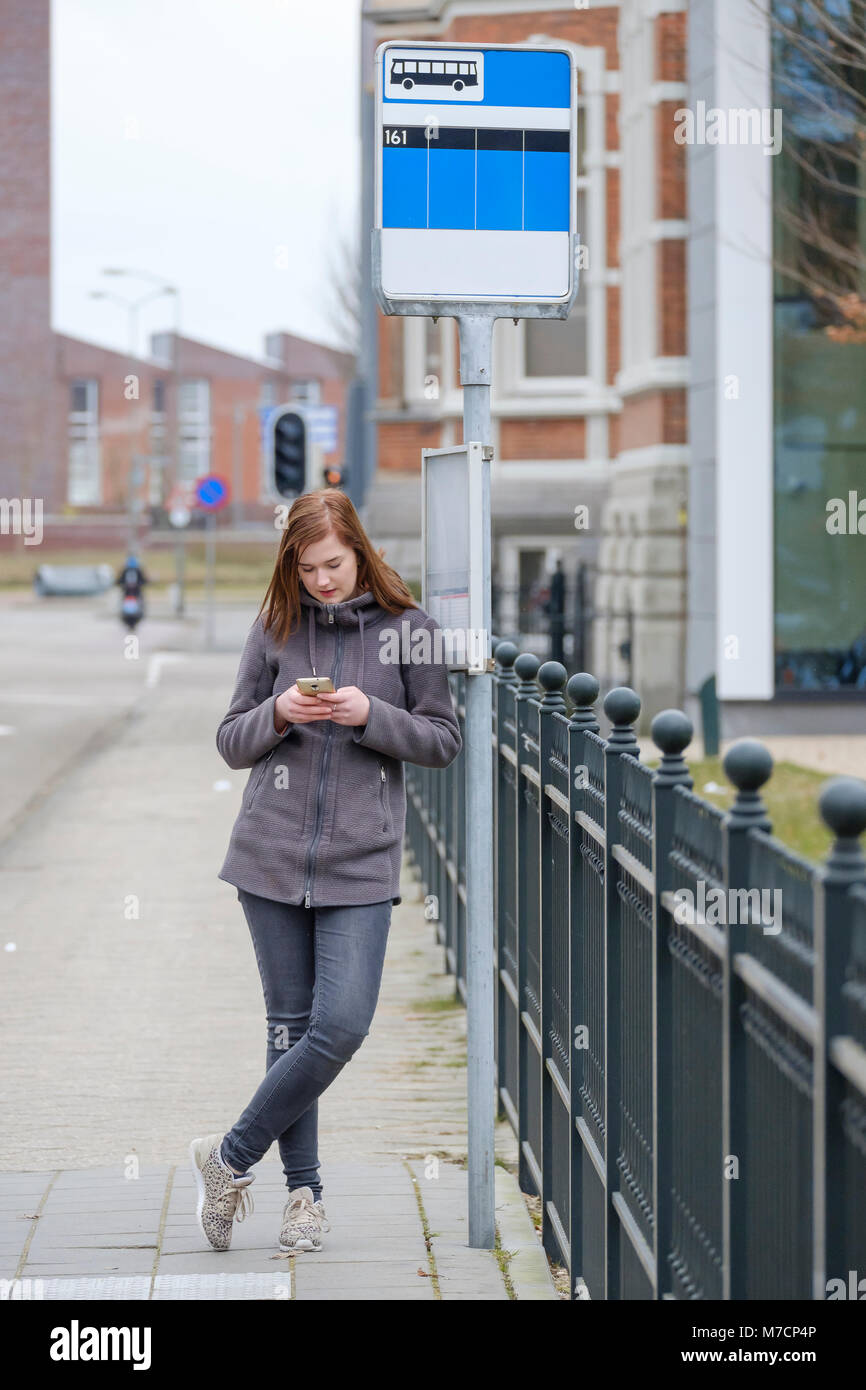 Junge Frau steht an einer Bushaltestelle und schauen, während sie darauf warten, Langeweile auf Ihrem Telefon Stockfoto