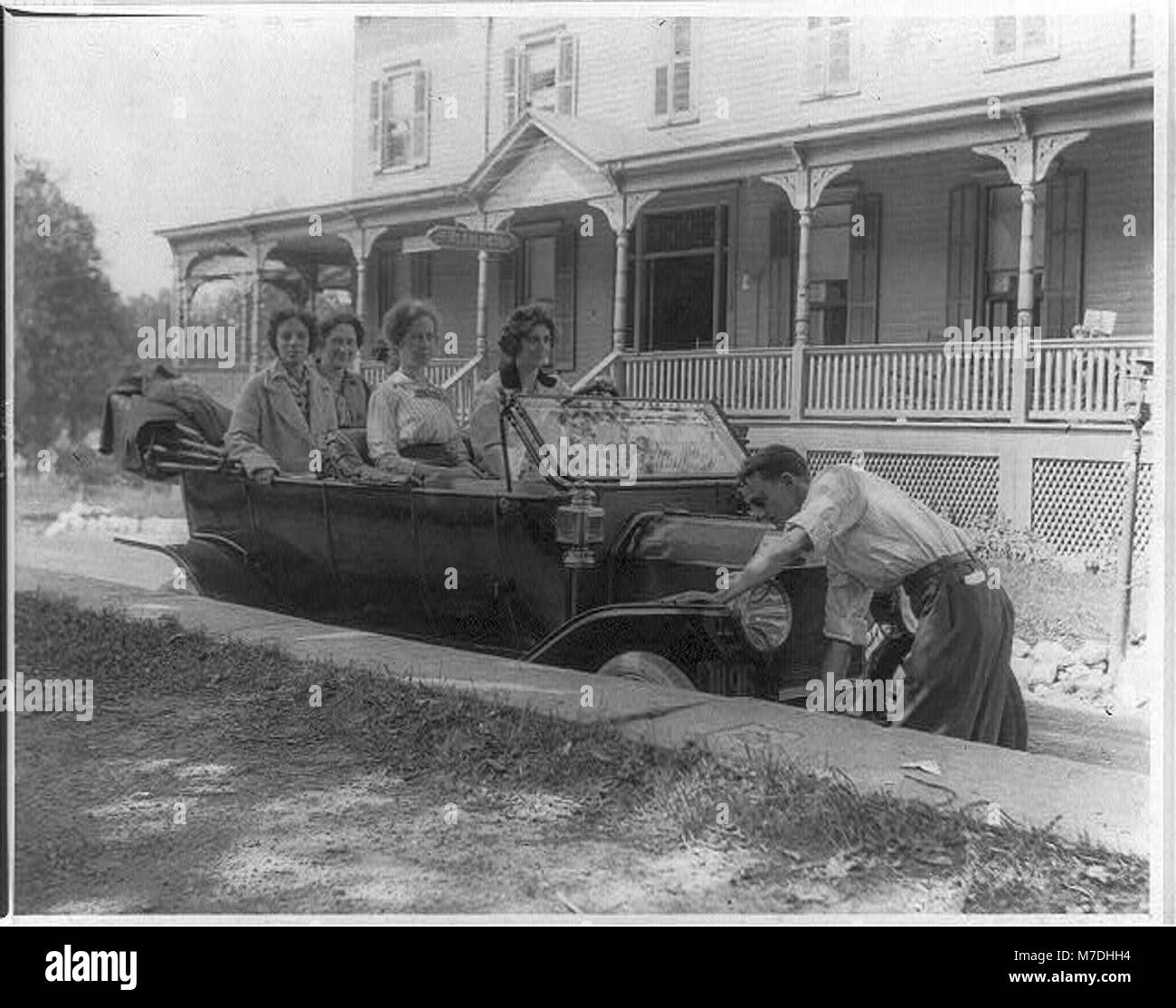 Ein Vintage-Foto, das einen Mann zeigt, der ein Auto vor dem Mt. Arlington Hotel, begleitet von fünf Frauen, spiegelt die frühe Automobilkultur wider. Stockfoto