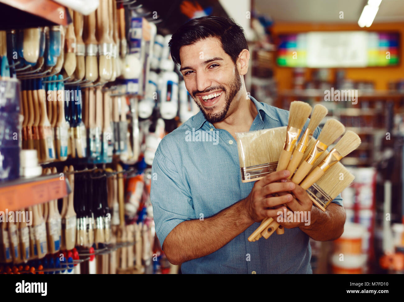 Junger Mann, der unter den Regalen in Farbe Speicher Auswahl von Pinseln Zufrieden Stockfoto