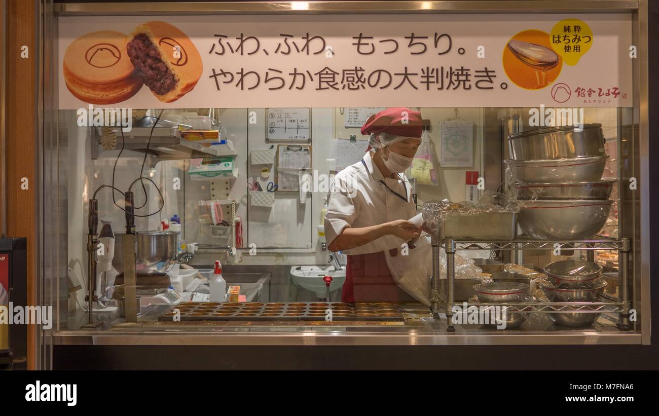 Japanische Bäcker Tragen von Red Hat und Schürze umgeben von Schalen, die traditionelle Japanische Rote Bohne Kuchen schoss durch Bäckerei Fenster. Stockfoto