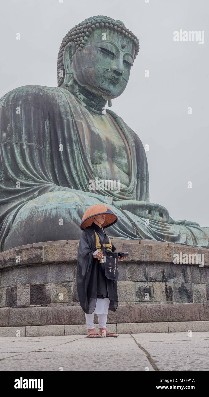 Japanischer buddhistischer Pilger in traditionellen Schüssel geformte Stroh Hut gekleidet, schwarze und weiße Roben stand vor der Kamakura Buddha läutenden Glocke Stockfoto
