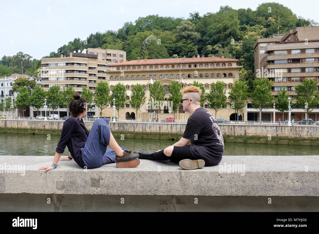 Junges Paar sitzt auf Wand, die mit dem Fluss Nervion, außerhalb des Guggenheim Museum, Bilbao, Vizcaya, País Vasco, Spanien, Stockfoto