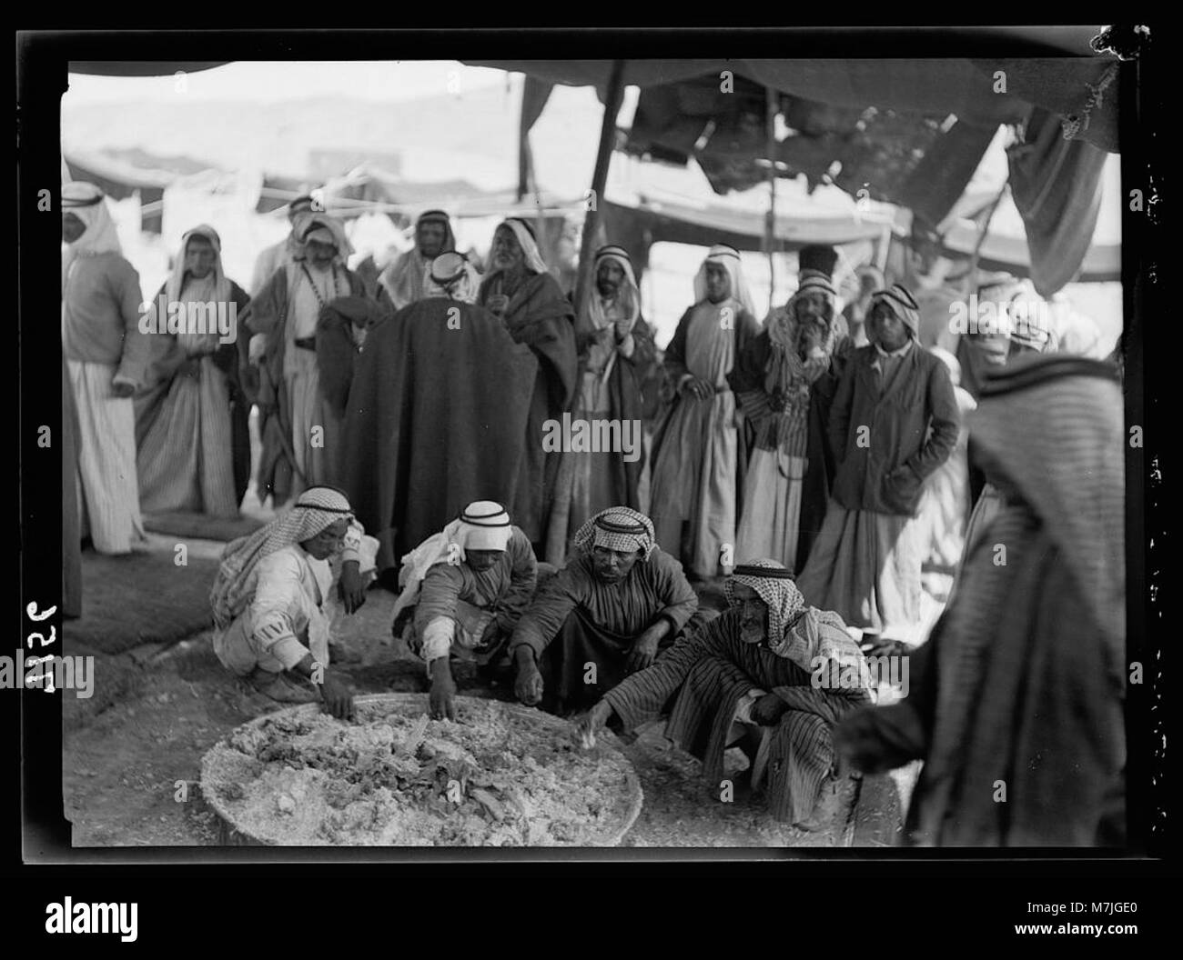 Beduinen versammeln sich um ein großes Tablett, um eine Mahlzeit am historischen Ort von Shunet Nimrin zu teilen. Das Foto zeigt eine traditionelle Beduinenspeise in einer Wüstenumgebung. Stockfoto
