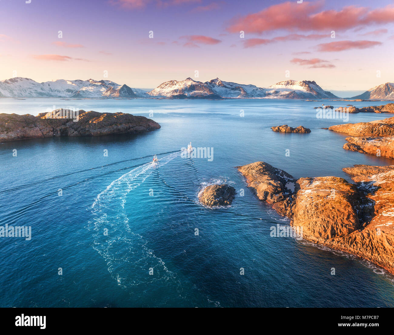 Luftaufnahme der Fischerboote, die Felsen in das blaue Meer, schneebedeckte Berge und bunte purple sky mit roten Wolken bei Sonnenuntergang im Winter auf den Lofoten Inseln, N Stockfoto