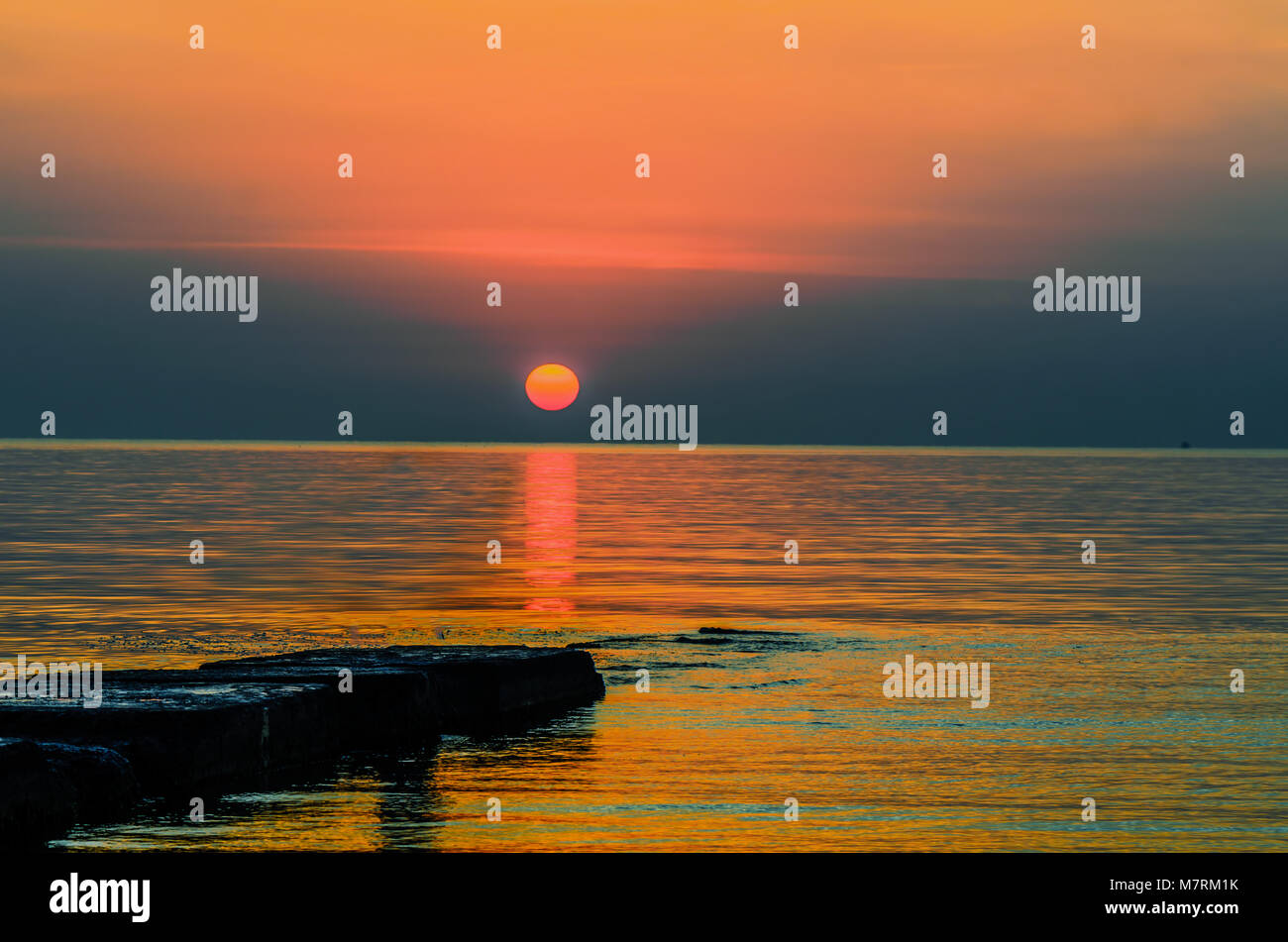 Orange rote Sonne über dem goldenen Wellen des Ozeans, die schwarzen Steine auf dem Stein Wasser, die Silhouette des Schiffes auf dem Blue Horizon Stockfoto