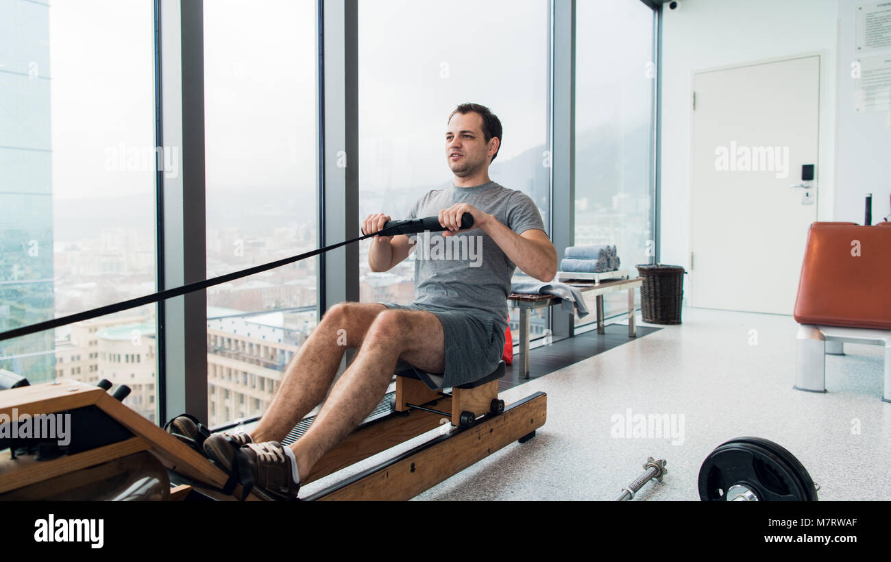 Rudern mit Strom. Seitenansicht des jungen Mannes in Sportkleidung, Rudern vor Fenster im Fitnessstudio. Stockfoto