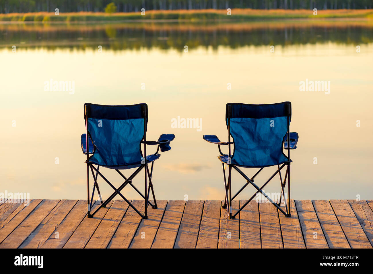 Ein paar leere Stühle stehen auf einer hölzernen Pier in der Nähe des Sees in der Morgendämmerung Stockfoto