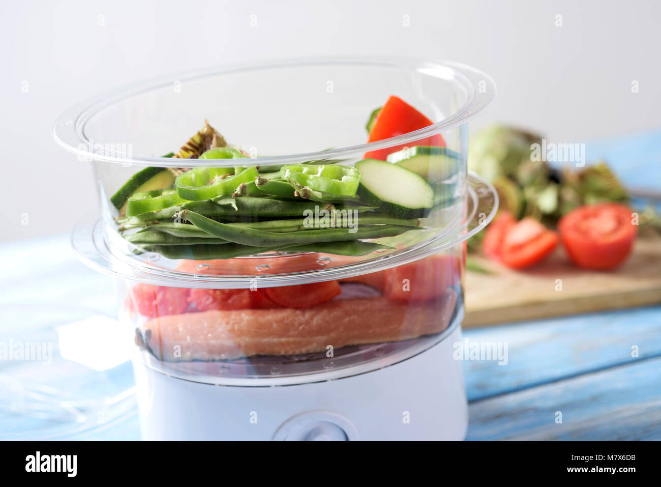 Nahaufnahme eines elektrischen Dampfgarer mit einigen rohen Lebensmitteln, wie grüne Bohnen, Zucchini, Paprika, Tomaten oder Fische, auf ein helles Blau Holztisch Stockfoto