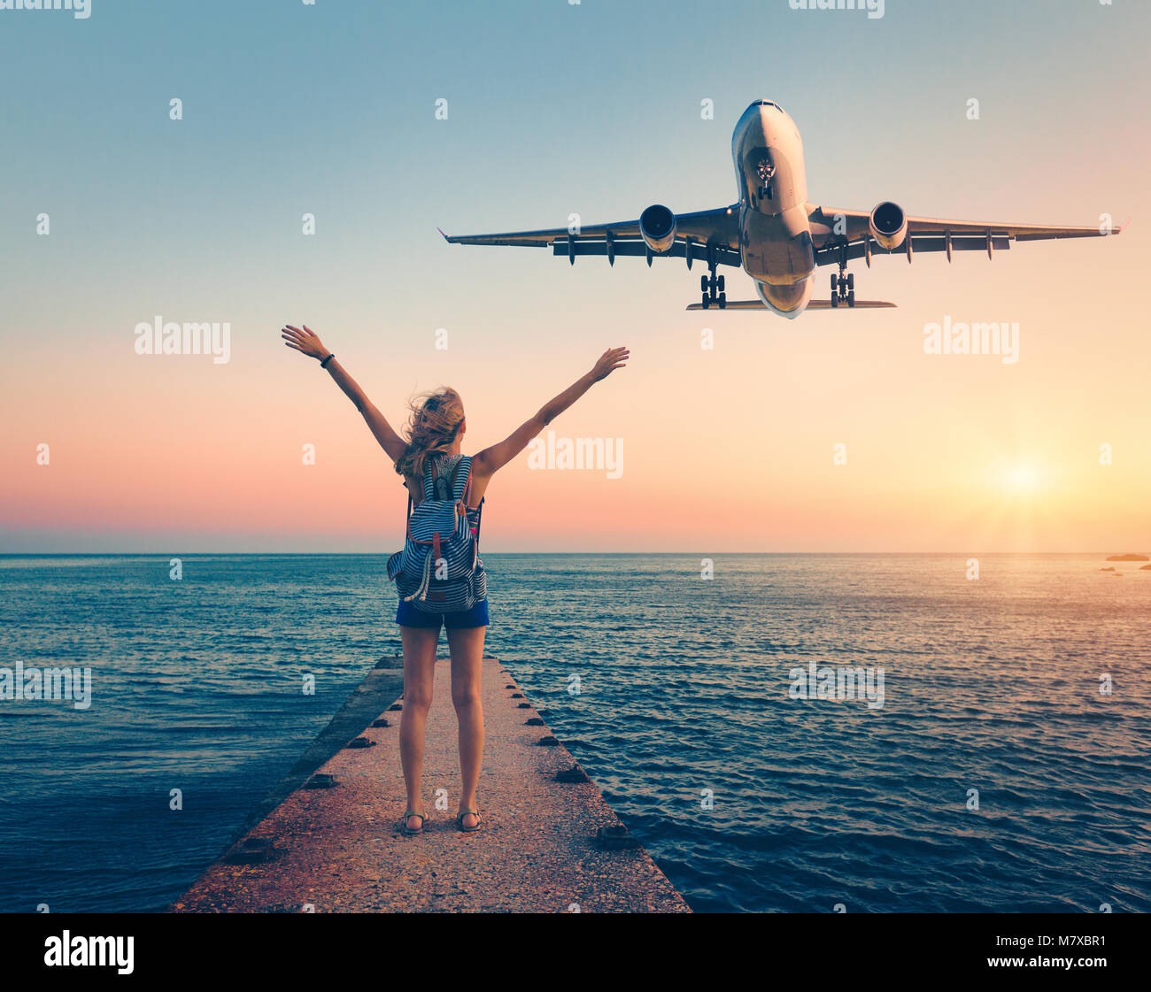 Flugzeug und Frau bei Sonnenuntergang. Sommer Landschaft mit Mädchen, der auf dem Meer Pier mit erhobenen Armen und fliegenden Passagierflugzeugs. Frau und Landung Stockfoto