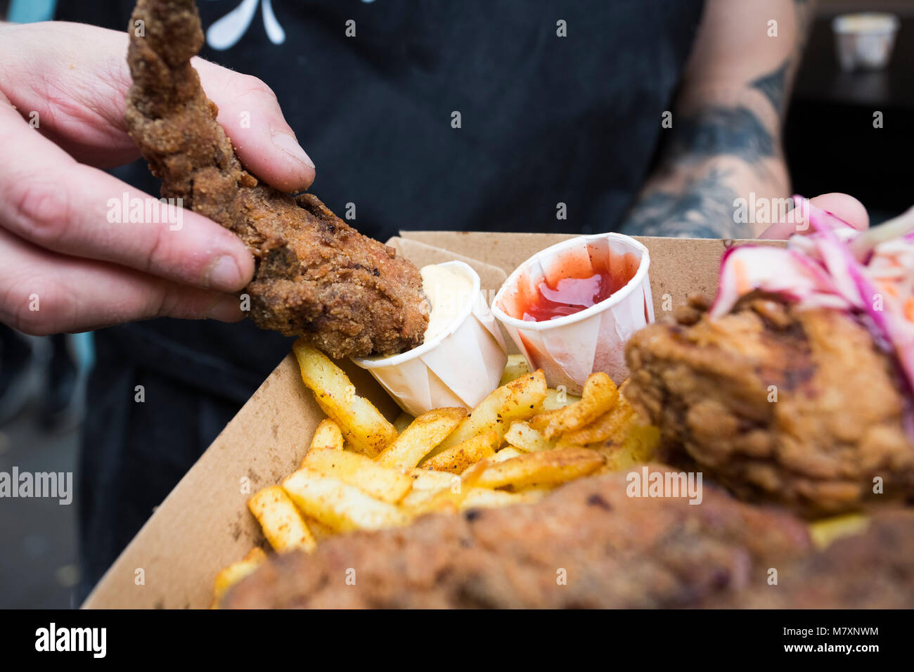 Fried Chicken und Chips mit Krautsalat, in Mayonnaise mit Ketchup getaucht vom Mann mit Tattoos. Hipster Street Food. Stockfoto