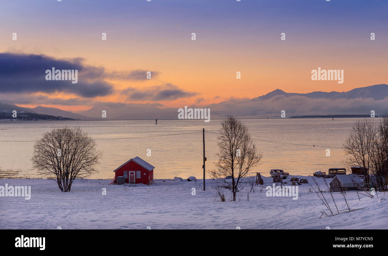 Eine Winterlandschaft in Nordnorwegen mit Schnee, Berge, Fjorde und einem roten Häuschen bei Sonnenuntergang. Stockfoto